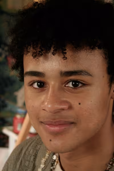 Headshot of a young person with short, curly dark hair looking directly at the camera with a slight smile. They are wearing a beaded necklace and a textured olive-green top. The portrait is warmly lit, with a softly blurred indoor background featuring colorful artwork and studio elements.