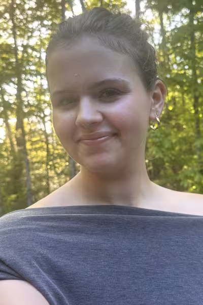 Outdoor headshot of a smiling young woman with dark hair pulled back, wearing small earrings and a septum ring. She is dressed in an off-the-shoulder gray top and looks directly at the camera. Sunlight filters through the trees behind her, creating a softly blurred forest backdrop.