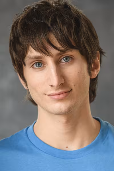Headshot of a young person with short dark brown hair and blue eyes, wearing a light blue crew-neck shirt and looking directly at the camera with a slight smile. The portrait is set against a neutral gray studio background.