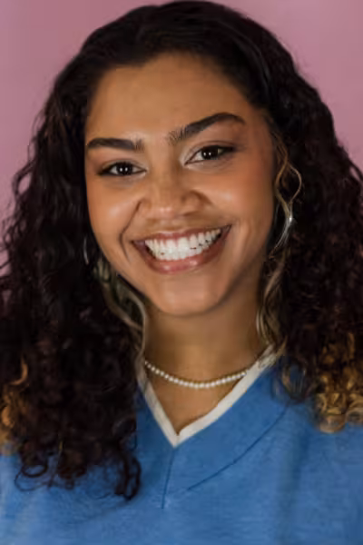 Head-and-shoulders portrait of a smiling young woman with dark curly hair featuring lighter highlights. She wears a light blue sleeveless top, hoop earrings, and a pearl necklace. The subject faces the camera with a bright smile against a soft pink studio background.