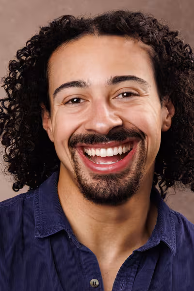 Head-and-shoulders portrait of a smiling man with shoulder-length dark curly hair, a mustache, and a goatee. He is wearing a dark blue collared shirt and looking directly at the camera. The photo is set against a soft brown studio background with even, natural lighting.
