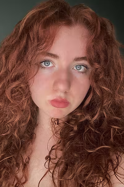Headshot of a young woman with long, curly red hair and blue eyes looking directly at the camera. She wears a small nose ring and has a neutral expression. The portrait is softly lit and set against a dark green studio background.
