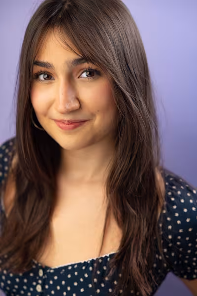 Head-and-shoulders portrait of a young woman with long dark brown hair and brown eyes, smiling softly at the camera. She wears a navy blue dress with a white floral pattern and hoop earrings. The photo is set against a soft lavender background with gentle studio lighting.