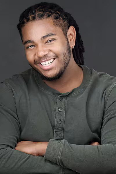 Headshot of a smiling young man with long braided hair pulled back, a trimmed beard, and a small hoop earring. He is wearing a dark green long-sleeve Henley shirt and is posed with his arms crossed while looking directly at the camera. The portrait is set against a neutral gray studio background.