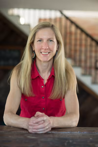 Portrait of a smiling Bonnie Jo Campbell with long blonde hair wearing a red sleeveless button-up shirt and patterned dangling earrings. She is seated behind a wooden table with her hands clasped, with a softly blurred wooden staircase and interior in the background.