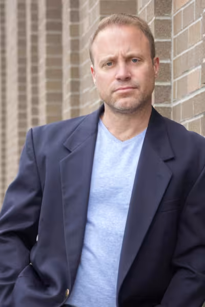 Chad Broughman wearing a navy blazer over a light blue V-neck shirt stands against a tan brick wall, looking directly at the camera with a neutral expression.