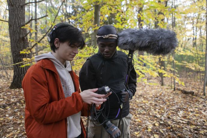 Two Film & New Media students, surrounded by forest, look at a camera's viewfinder.