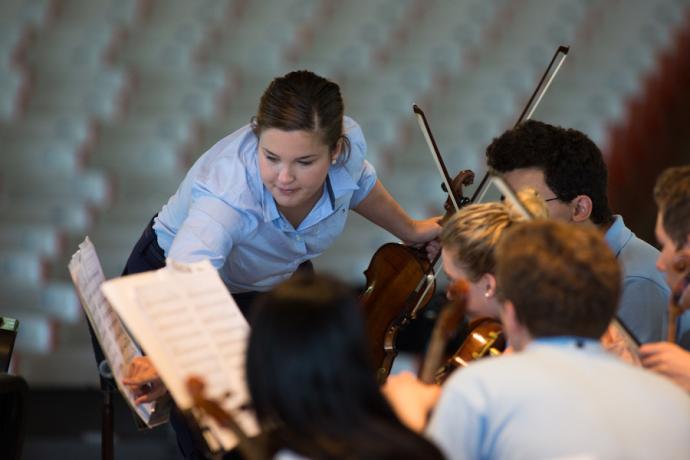 A woman in a blue polo shirt leans over to point at a sheet of music. She is surrounded by young violinists in pale blue polo shirts.