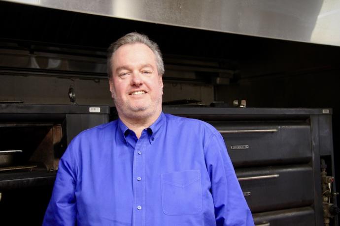 A man in a blue shirt stands in front of a large industrial oven and smiles.