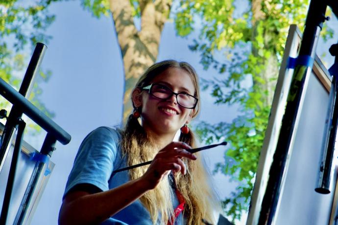 A young girl with glasses and long blonde ponytails looks up from her painting to smile.