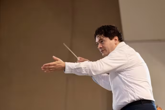 A dark-haired man wearing a white shirt leans forward to conduct an orchestra.