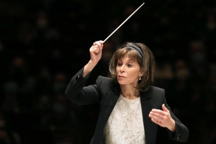 A woman wearing a black jacket and white shirt waves a conductor's baton in the air.
