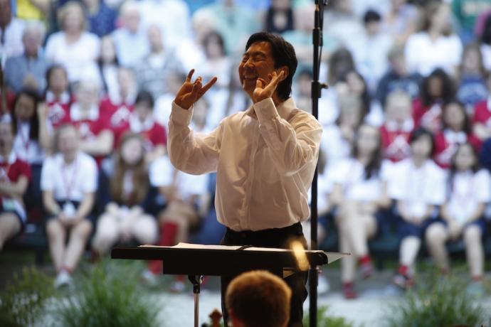 A conductor wearing a white shirt raises his hands and smiles at his orchestra.