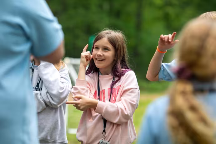 A student wearing a pink shirt smiles and lifts her arms as if dancing.