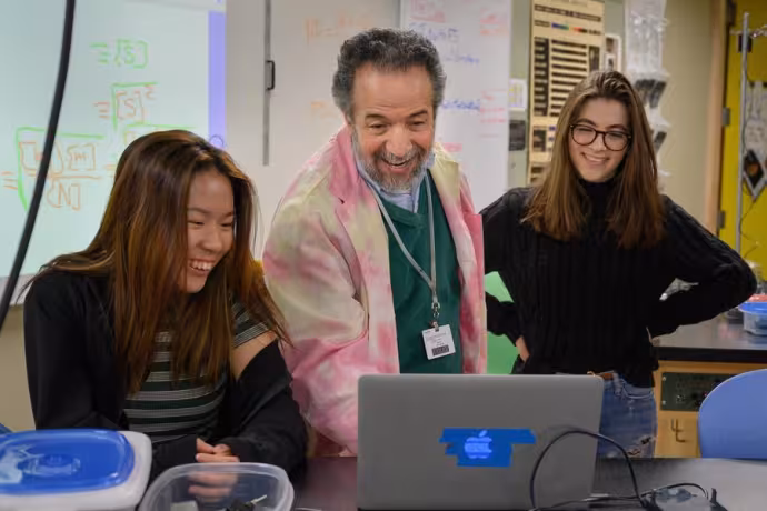 A teacher dressed in a brightly-colored coat smiles as he helps two students with their classwork.