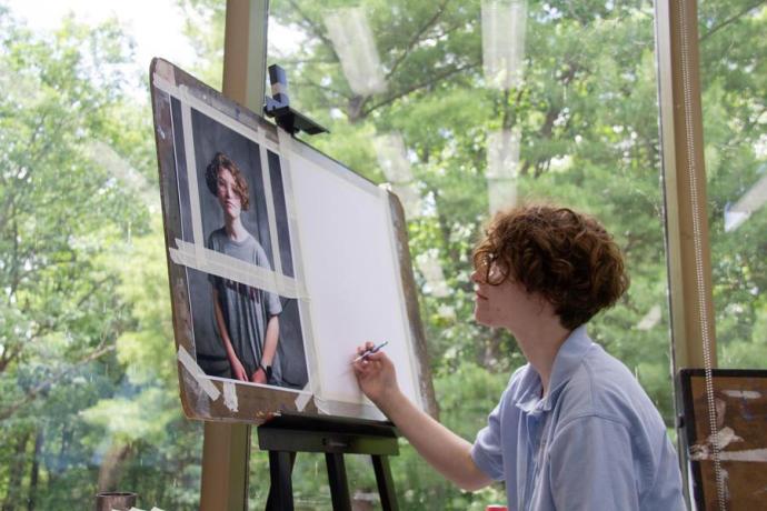 A student in a light blue shirt works on a self-portrait beside a window looking out into the forest.