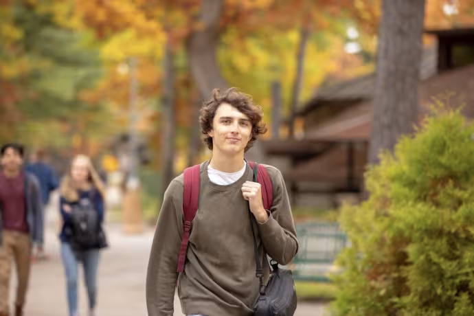 A smiling student wearing a green shirt strides down a sidewalk on Interlochen's campus.