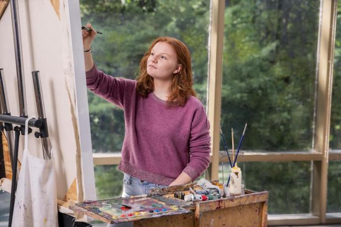 A visual arts student with long red hair works on a painting. 