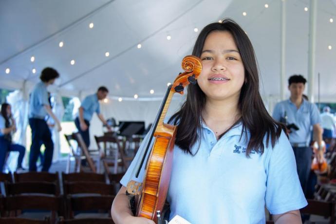 A young musician in a pale blue polo smiles as she holds her violin. 