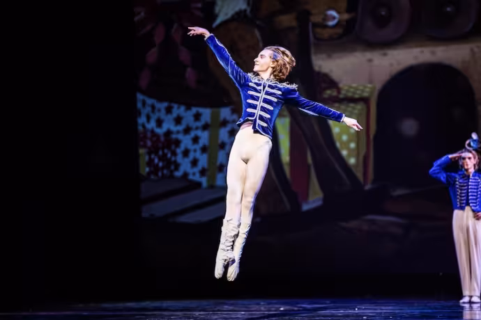 A dancer dressed in a blue jacket and white tights leaps into the air.