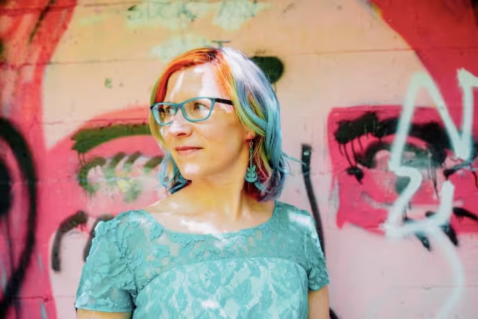 A woman with brightly colored hair stands in front of a graffiti background.