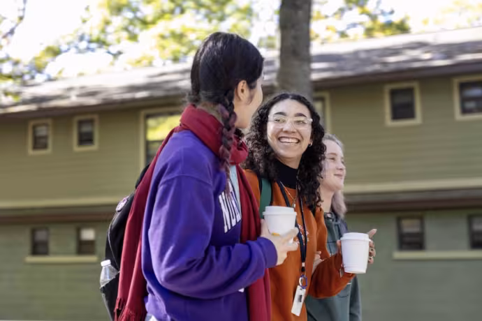 Smiling students stroll on campus with coffees in their hands.