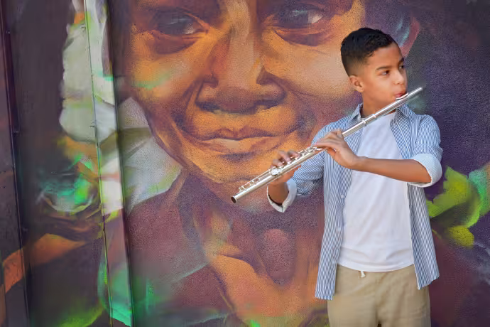A young boy plays his flute in front of a mural of a woman's face.