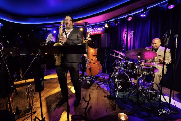 A saxophonist dressed in a dark suit plays passionately in front of a neon-lit stage with a drummer.