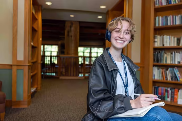 A student wearing headphones and holding an open book in their lap smiles at the camera.