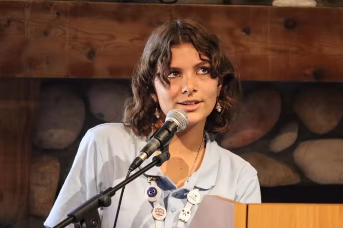 A creative writing student stands at a podium with a microphone, reading from a piece of paper.