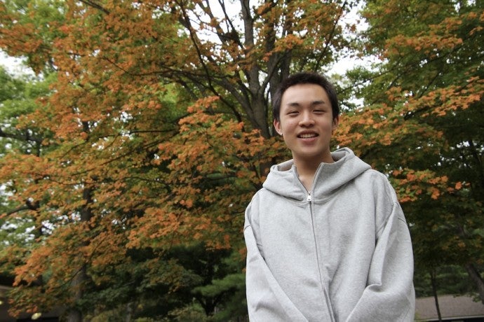 A student in a white hoodie stands against a fall landscape.