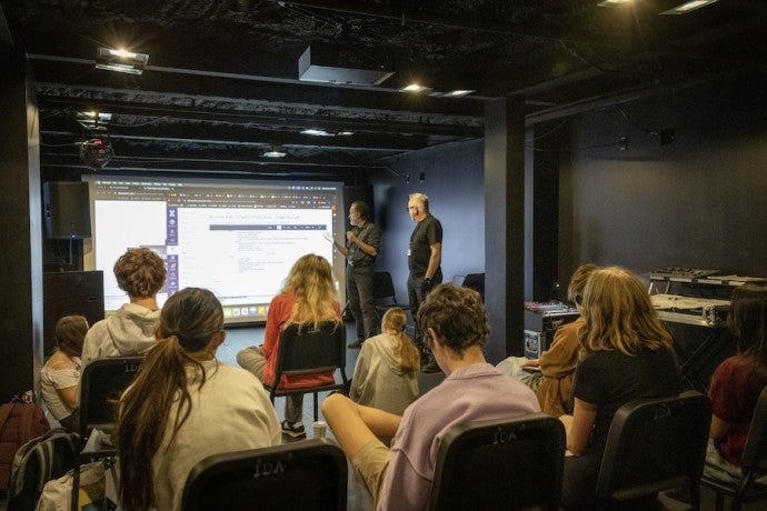 Two instructors gesture towards a screen in front of a group of seated students.