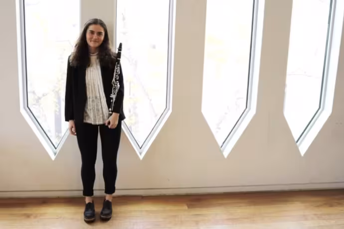 Katia Waxman stands in front of a row of windows. She is dressed professionally and holding her clarinet.