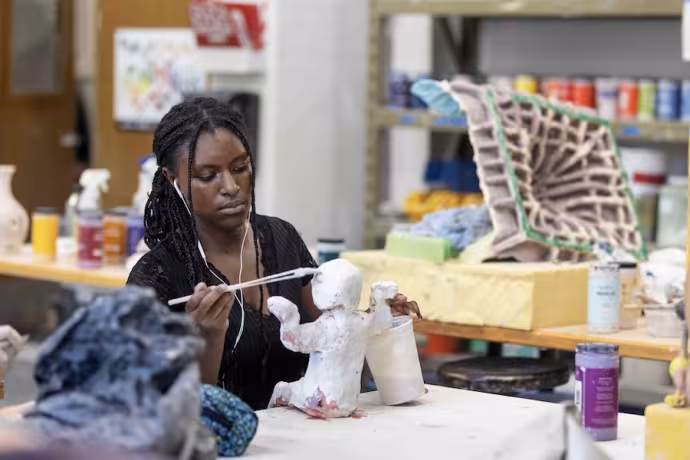 A seated student wearing earbuds paints a small white figure.