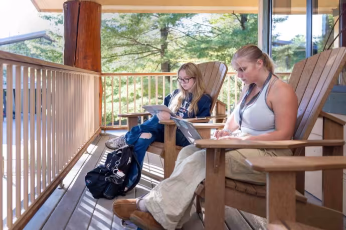 Two students sit comfortably on a deck as they type on their laptops.