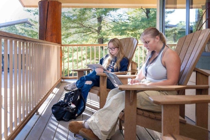 Two students sit comfortably on a deck as they type on their laptops.
