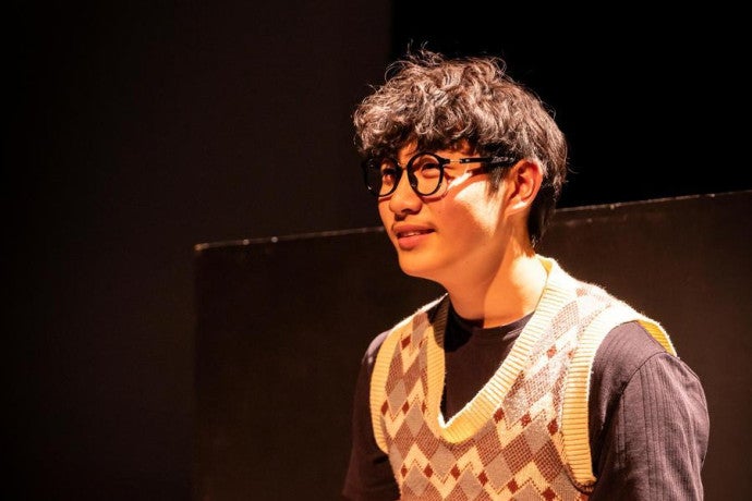 A student wearing an argyle vest sits in dramatic stage lighting.