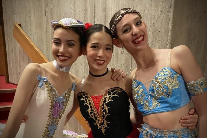 Three student dancers in colorful costumes hug each other and smile.