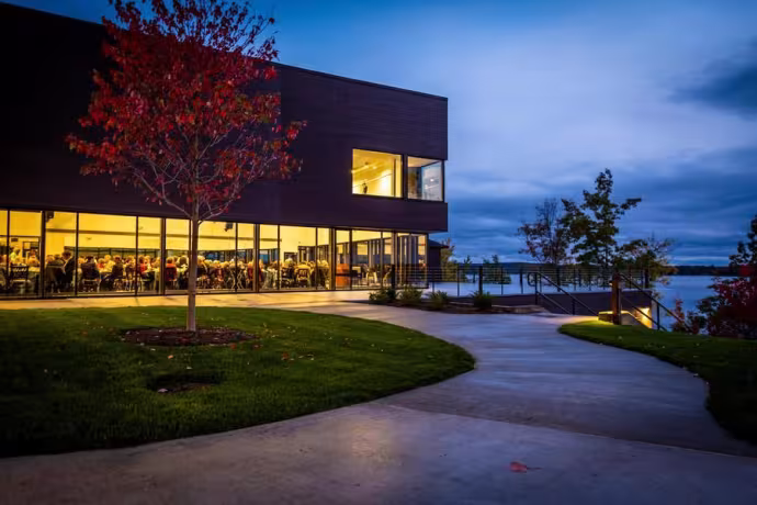 A large building with warmly-lit studio windows overlooks a shoreline at dusk.