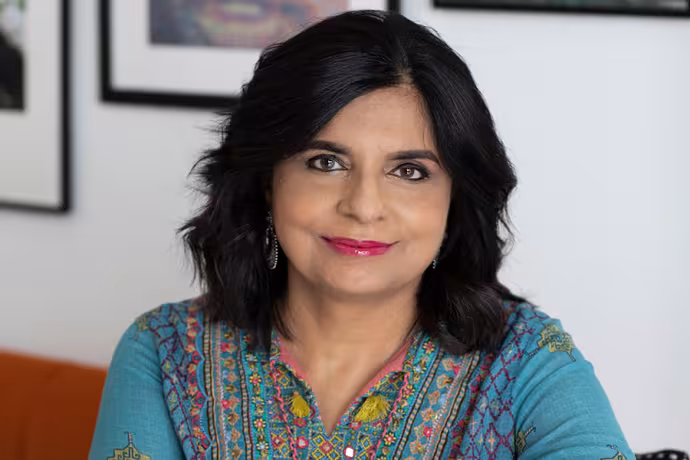 Professional headshot of a Parul Kapur with shoulder-length dark hair, wearing a teal embroidered top and earrings, smiling gently while facing the camera against a softly blurred indoor background with framed artwork.