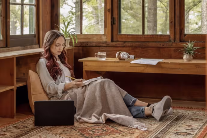 A young writer sits comfortably on the floor with a laptop and notebook.