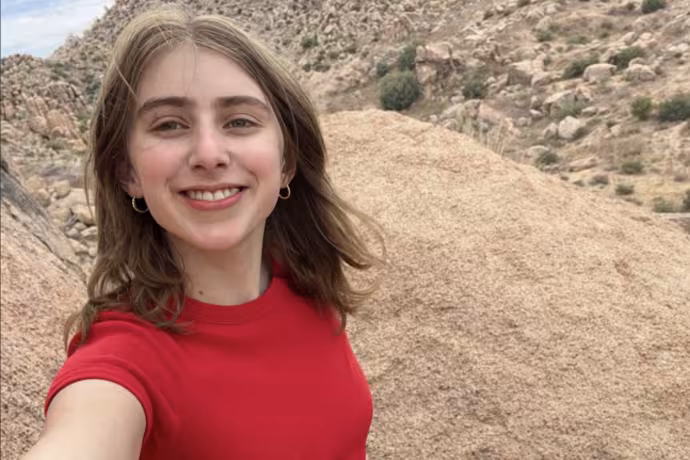 A young woman in a red shirt with a jubilant expression stands at the top of a mountain.