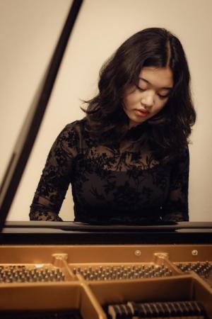 A dark-haired student dressed in black lace bends over a piano as she plays it.
