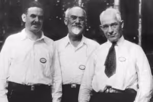 A vintage black and white photo of three men in white shirts and name tags standing outdoors.