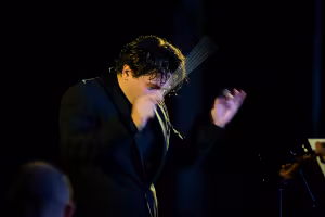 Cristian Măcelaru conducts passionately on a dimly lit stage, raising his baton with expressive motion while wearing a black suit.