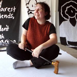 Oaklee Thiele, a petite dark-haired woman, smiles as she sits cross-legged in front of several large, black-and-white banners.