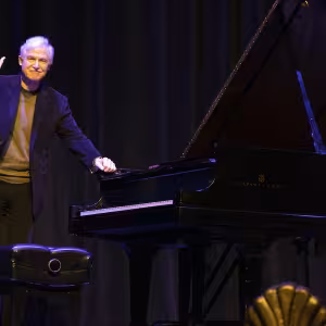 A grey-haired man in his 70, standing next to a black grad piano, waves to an audience following his performance.