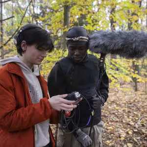 Two Film & New Media students, surrounded by forest, look at a camera's viewfinder.
