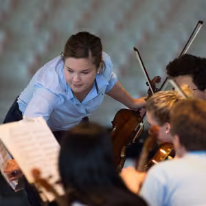 A woman in a blue polo shirt leans over to point at a sheet of music. She is surrounded by young violinists in pale blue polo shirts.