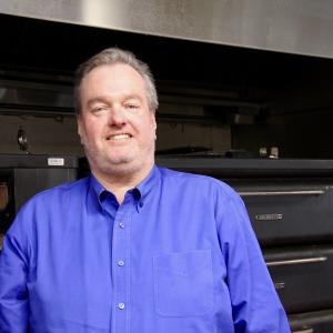 A man in a blue shirt stands in front of a large industrial oven and smiles.